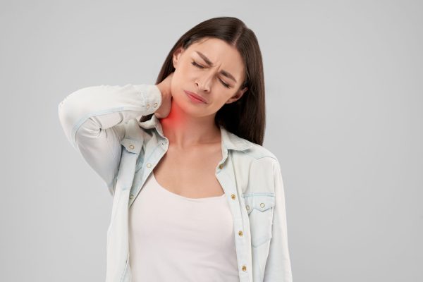 Young caucasian woman in white shirt over gray isolated background touching neck with hand with painful expression because of ache. Front view of brunette with closed eyes.